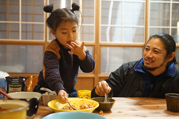 女の子がスプーンでご飯を食べ、その様子を父親が笑顔で眺めている写真。