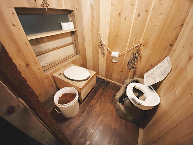 木でつくられたトイレ小屋の内部写真。便器の横におがくずのはいったバケツが置かれてある。