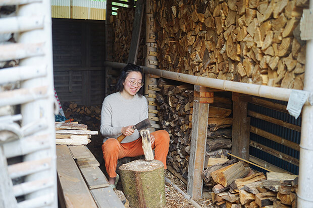 たくさんの薪が積み上げられた薪小屋で、薪を割る女性の写真。